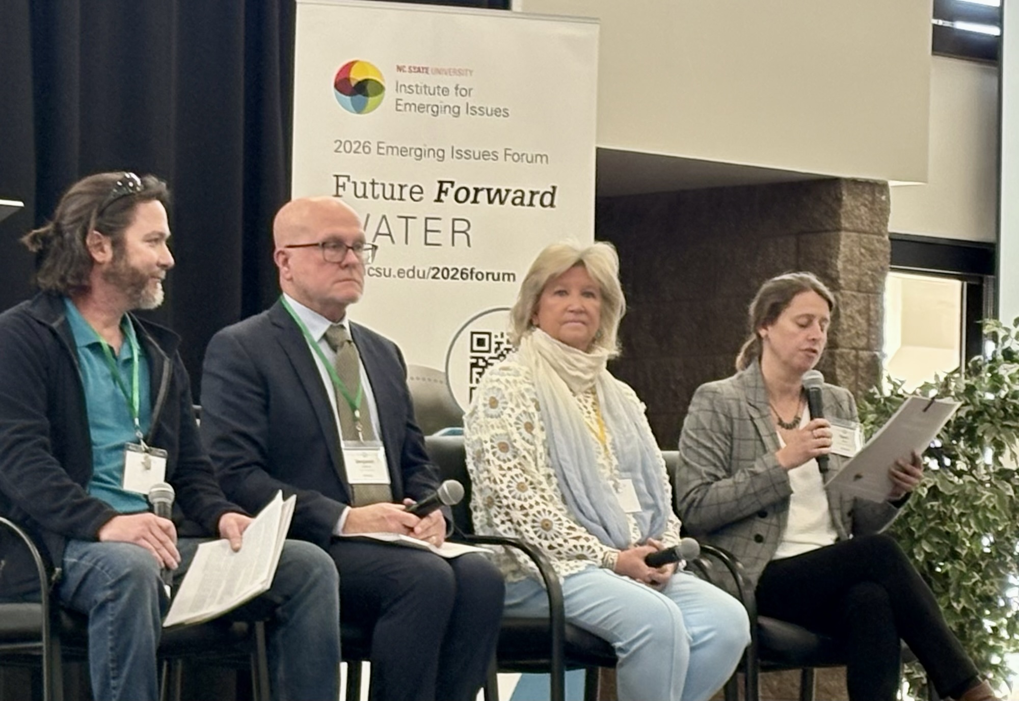 four people sit on a stage as one person on the right speaks into a microphone.